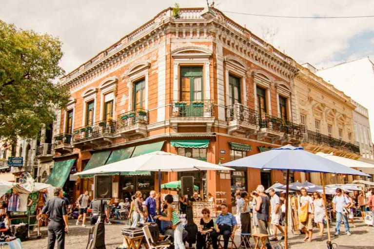 San Telmo Una esquina en san telmo con un edificio naranja ornamentado con balcones, rodeado de gente, puestos de mercado y sombrillas.