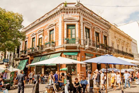 Una esquina en san telmo con un edificio naranja ornamentado con balcones, rodeado de gente, puestos de mercado y sombrillas.