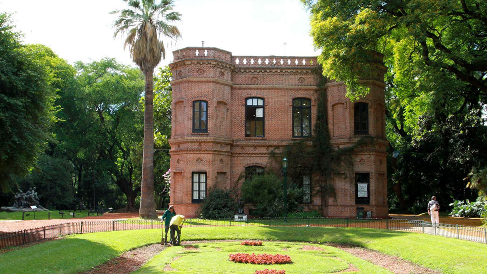 Camino a través de un jardín de rosas con gente sentada en bancos. Rosas rosas y rojas, árboles y un cielo azul.