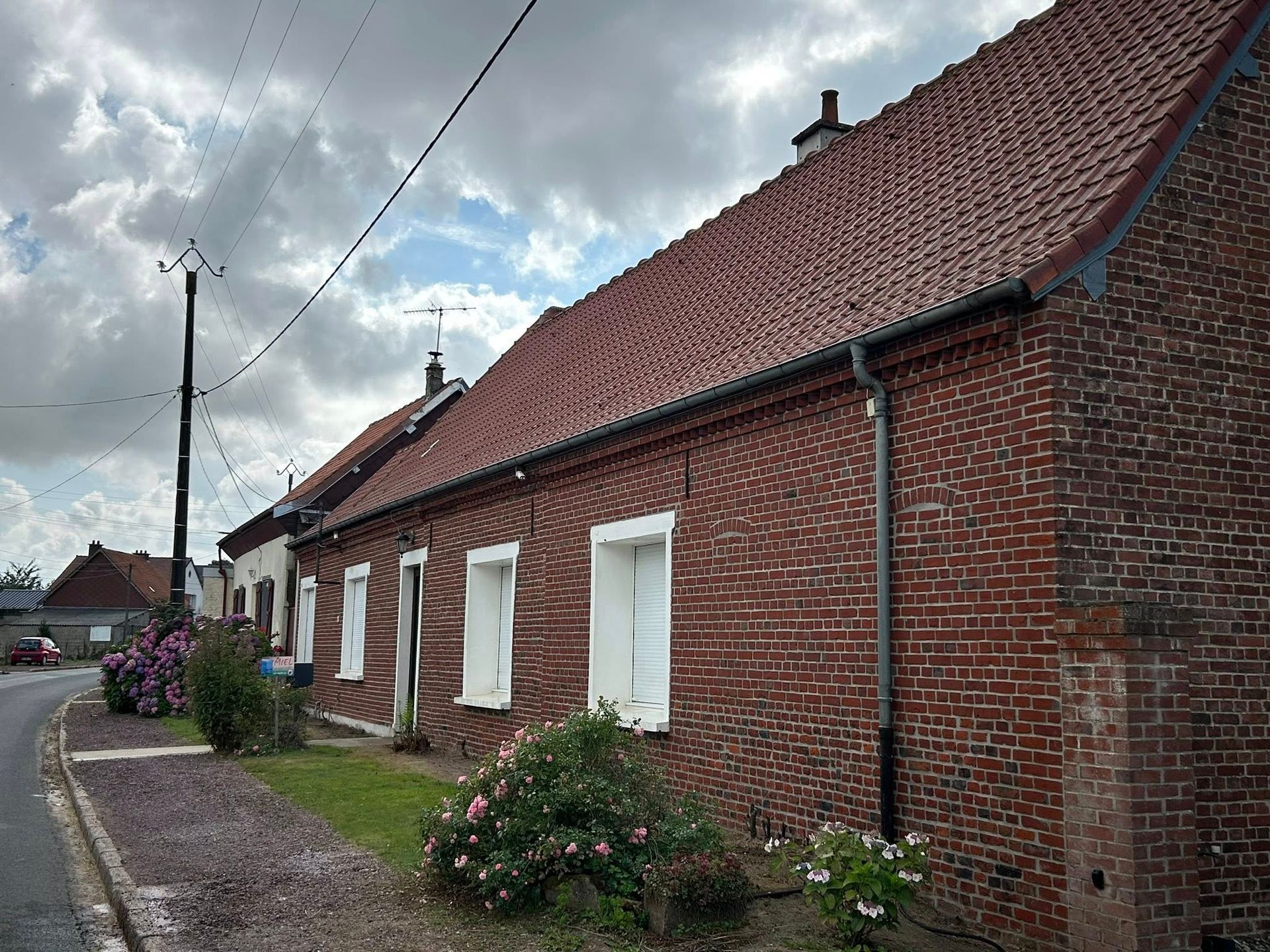 Des maisons en briques aux toits de tuiles rouges et aux volets blancs bordent une rue, sous un ciel nuageux.