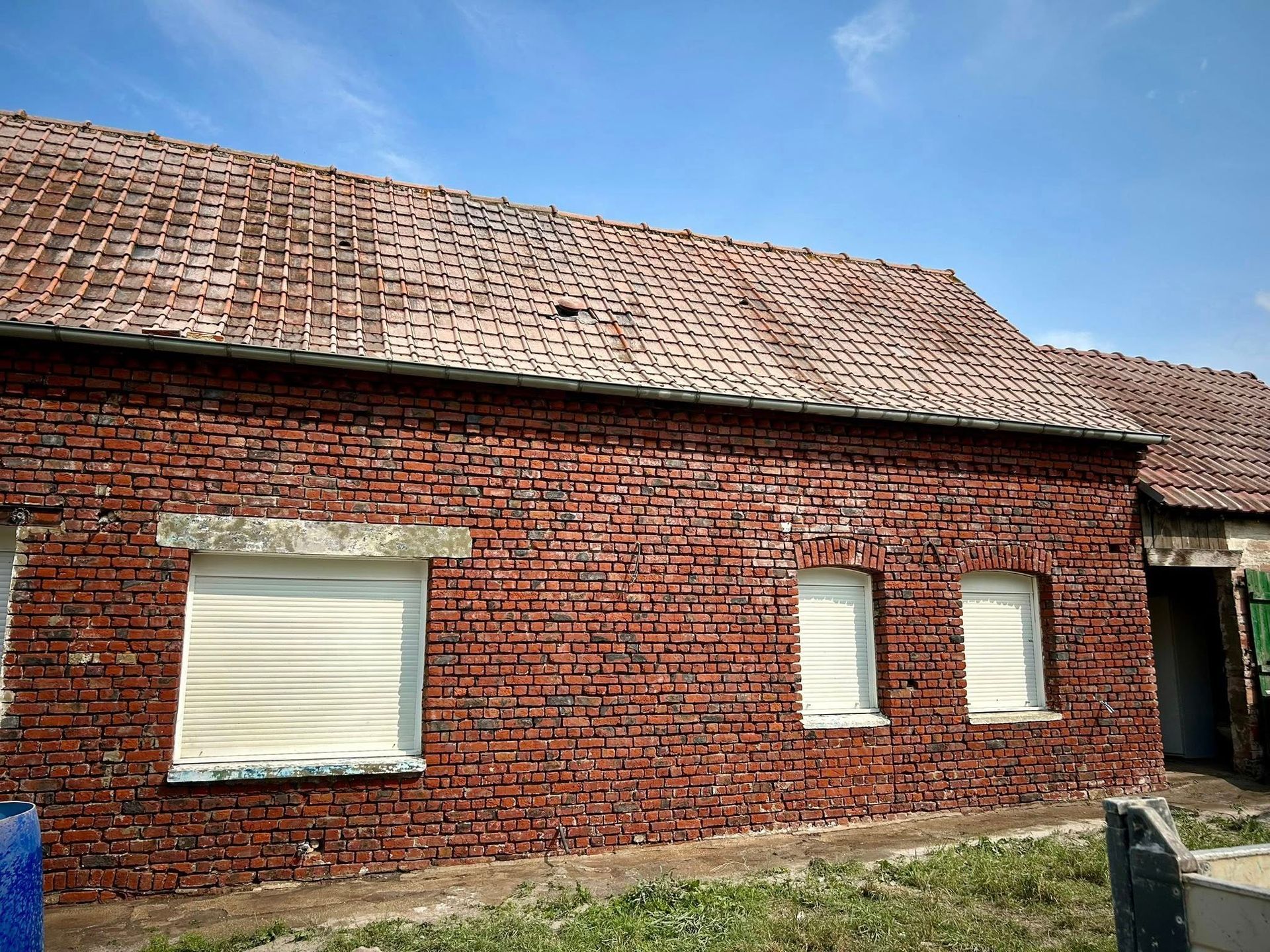 Bâtiment en briques avec un toit de tuiles rouges et trois fenêtres avec des volets fermés sur fond de ciel bleu.