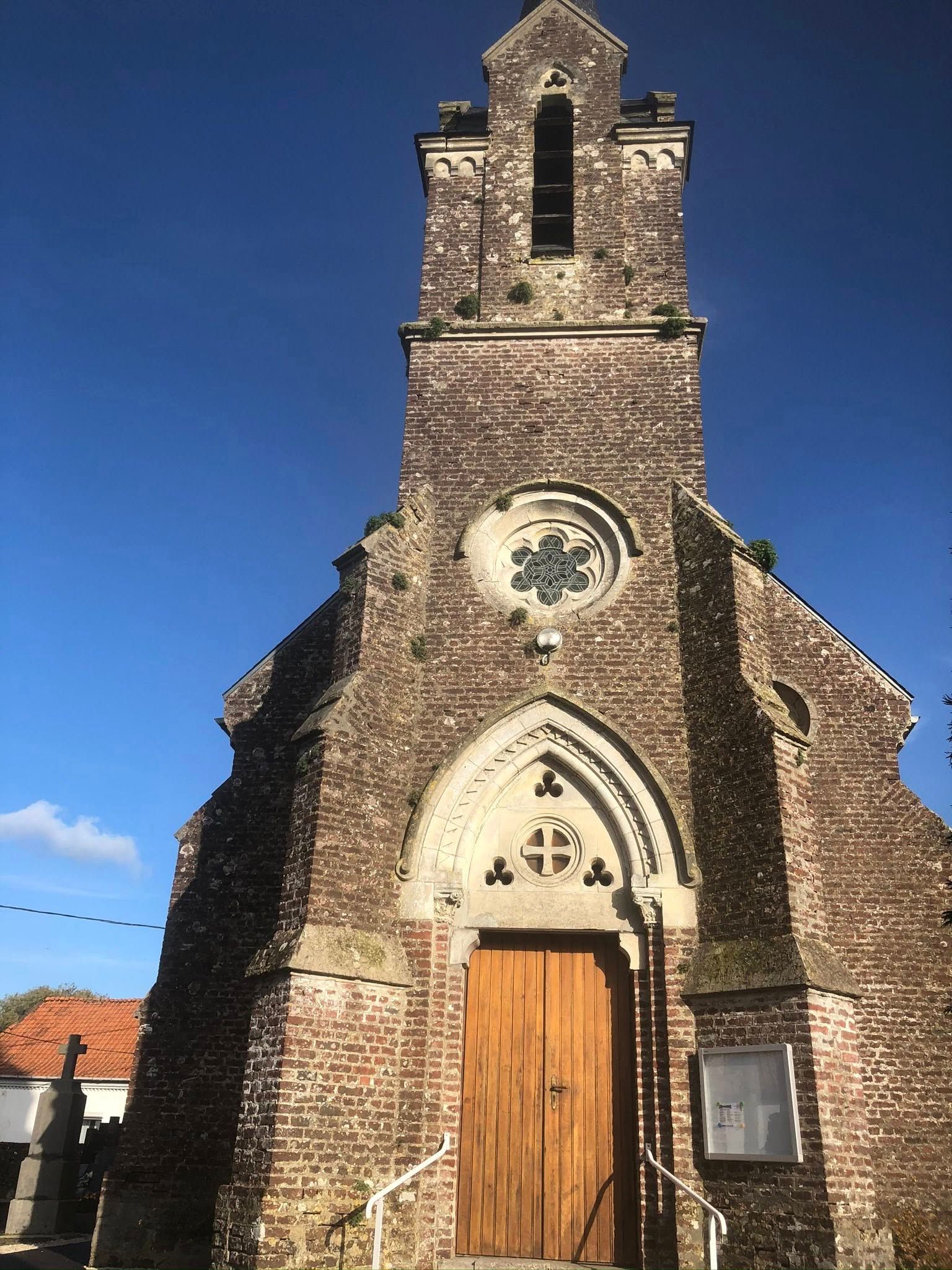 Église en briques avec un haut clocher, une porte en bois et une rosace sous un ciel bleu limpide.