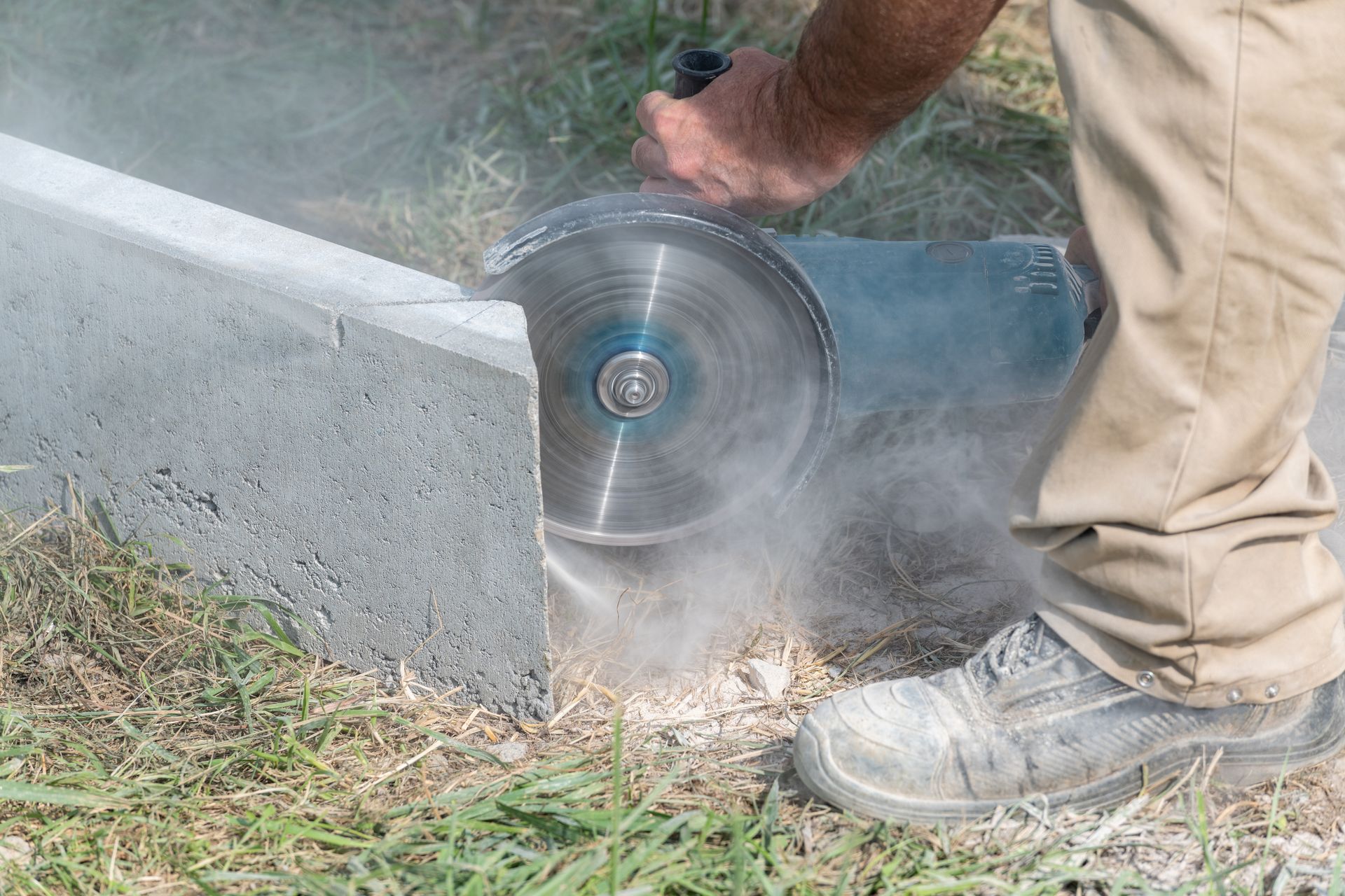 Une personne en pantalon beige découpe du béton à l'aide d'une scie électrique à l'extérieur, créant de la poussière.