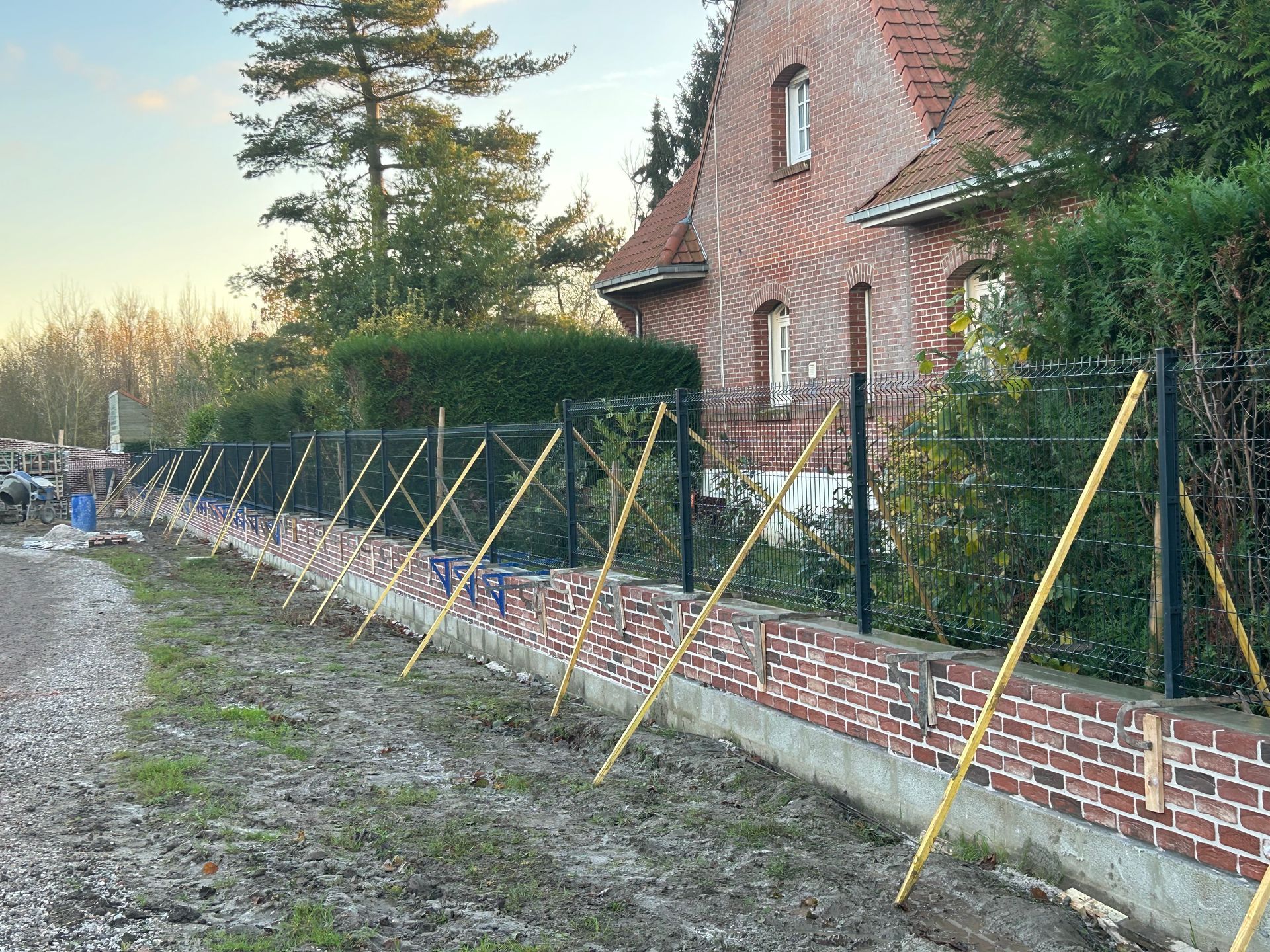 Un mur de briques avec une clôture noire en construction, soutenue par des étais jaunes. 