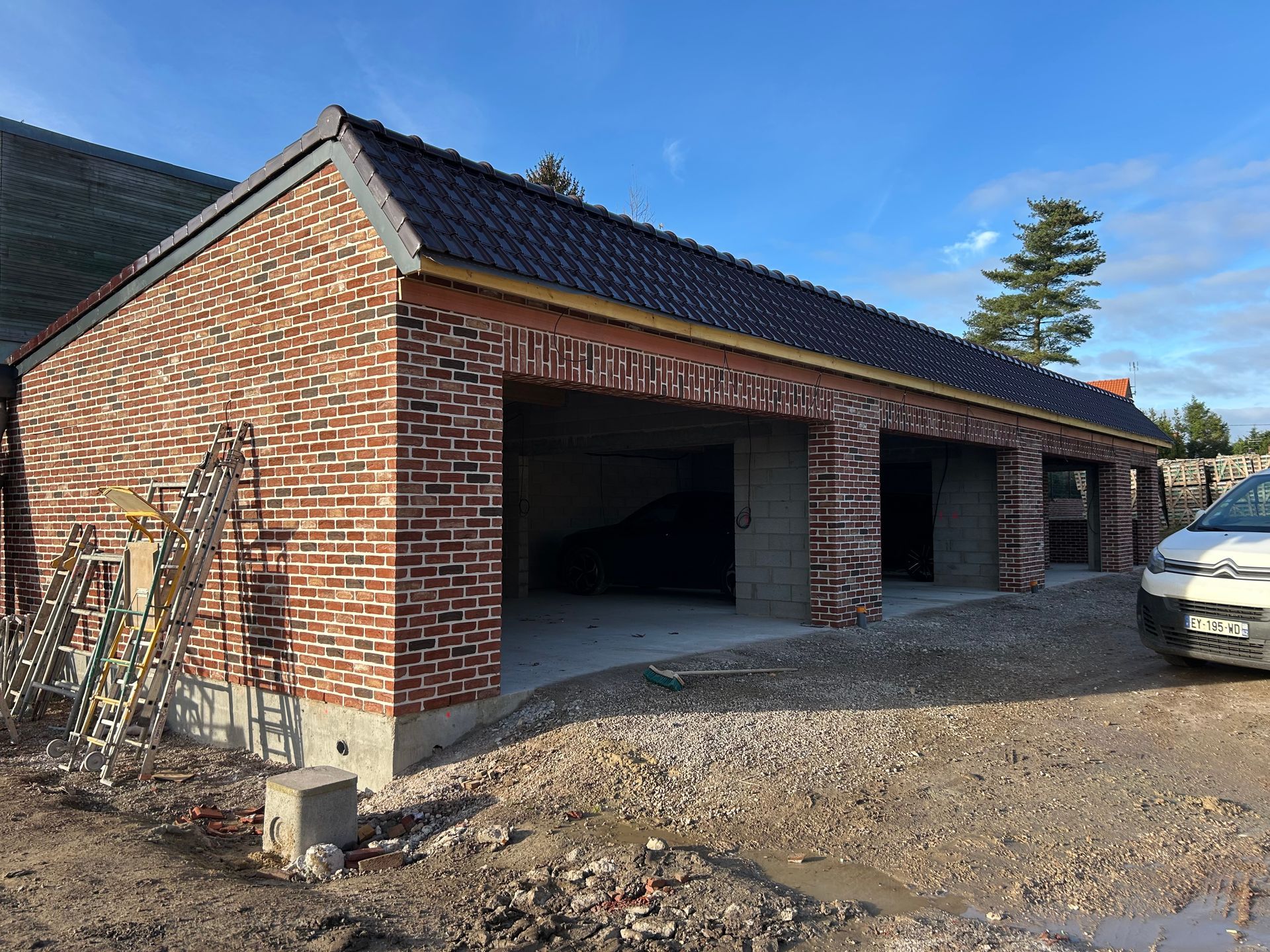 Garage en briques en construction avec deux travées, allée en gravier et ciel partiellement nuageux.