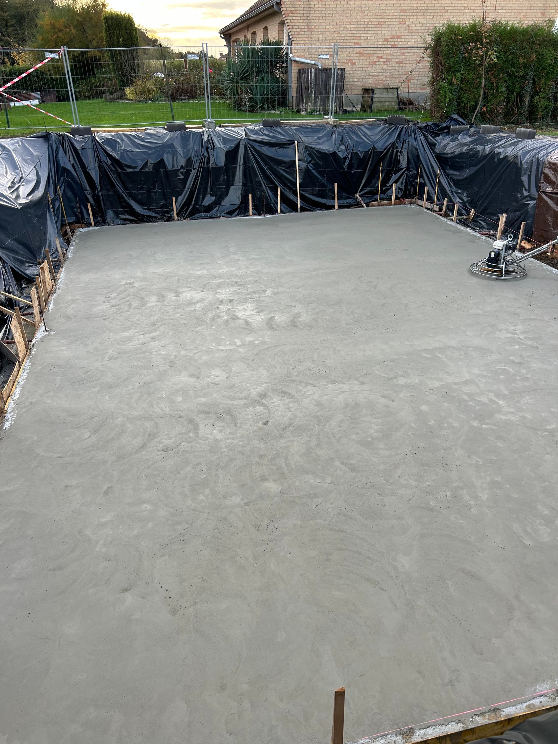 Une dalle de béton fraîchement coulée sur un chantier, entourée de bâches en plastique noir et de supports en bois.