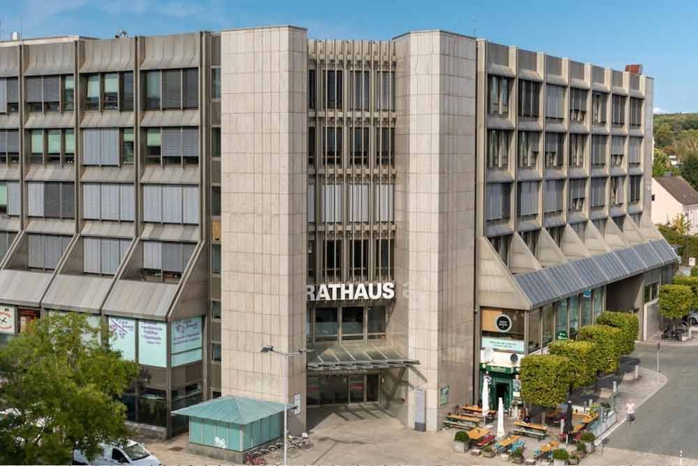 Rathausgebäude mit moderner Fassade und „RATHAUS“-Schriftzug; davor Sitzgelegenheiten im Freien und Bäume.