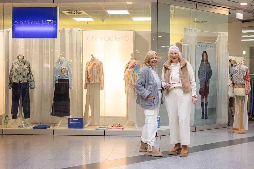 Zwei Frauen posieren vor einem Schaufenster eines Bekleidungsgeschäfts. Die eine trägt eine weiße Hose, die andere eine blaue Jacke.