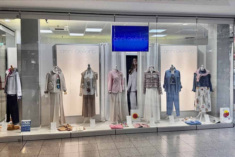 Schaufensterdekoration eines Bekleidungsgeschäfts mit Schaufensterpuppen in verschiedenen Outfits; neutrale Farben, in der Mitte hängt ein blauer Bildschirm.