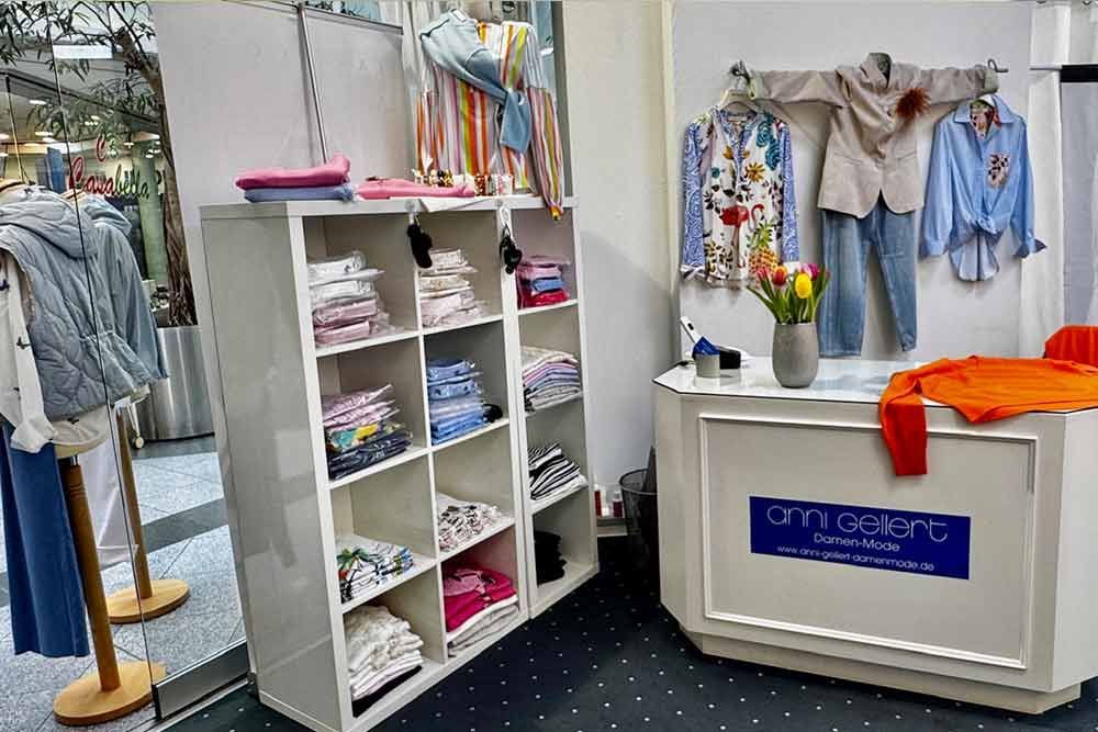 Kleiderauslage in einem Geschäft. Regale mit gefalteten Hemden. Hemden und Hosen hängen an Kleiderstangen. Weißer Tresen.