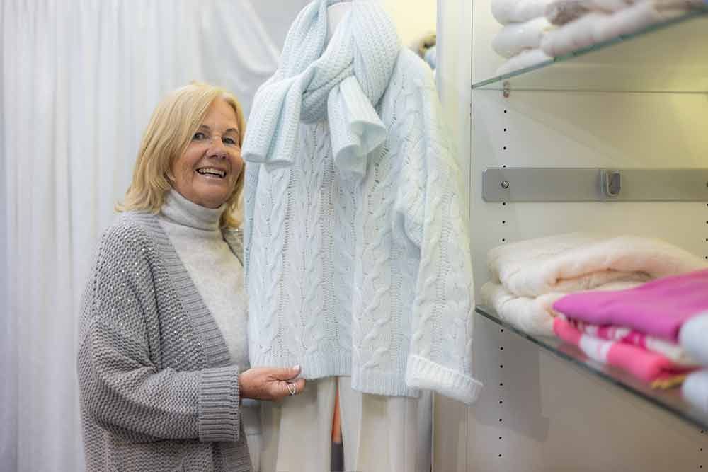 Eine Frau in einem Geschäft lächelt und hält einen hellblauen Pullover in der Hand. Ein passender Schal hängt daran.