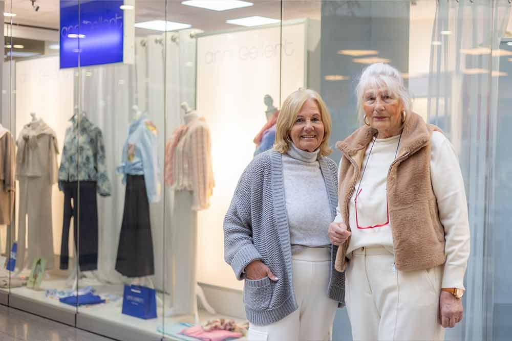 Zwei Frauen vor einem Schaufenster eines Bekleidungsgeschäfts. Die eine trägt einen grauen Pullover, die andere eine hellbraune Weste.