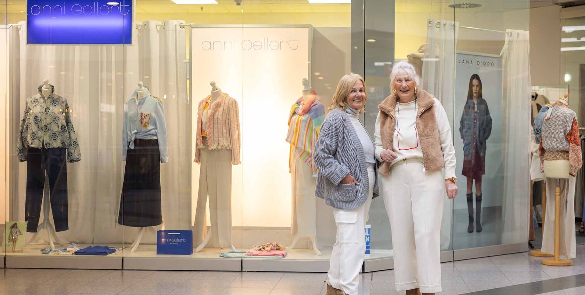 Zwei Frauen stehen vor einem Schaufenster. Die eine trägt einen hellblauen Pullover, die andere ein weißes Outfit. Im Schaufenster sind Schaufensterpuppen ausgestellt.