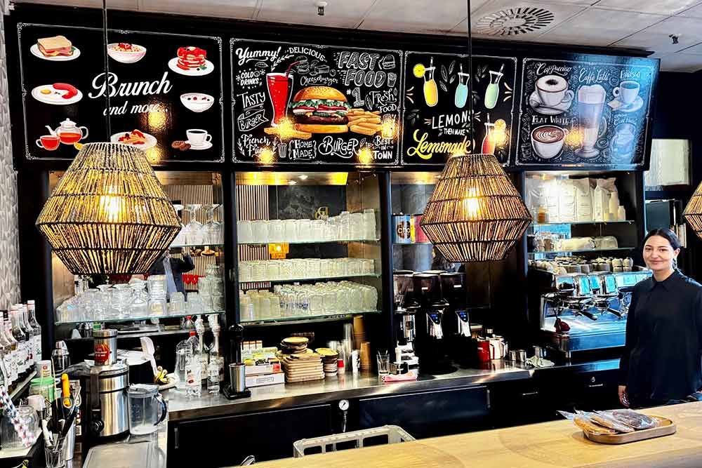 Eine Frau steht hinter der Theke eines Restaurants. Darüber hängen Lampen und Kreidetafeln mit Speisekarten.