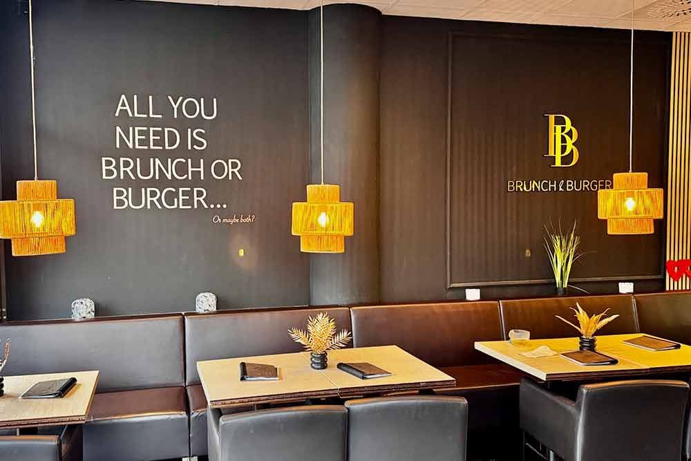 Restaurantinterieur mit Sitzgelegenheiten, gelben Pendelleuchten und einer Wand mit der Aufschrift „Alles, was du brauchst, ist Brunch oder Burger“.