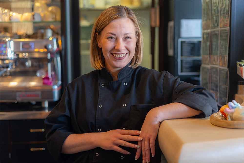 Eine Frau in schwarzer Kochjacke lächelt, während sie sich in einem Café an einen Tresen lehnt.