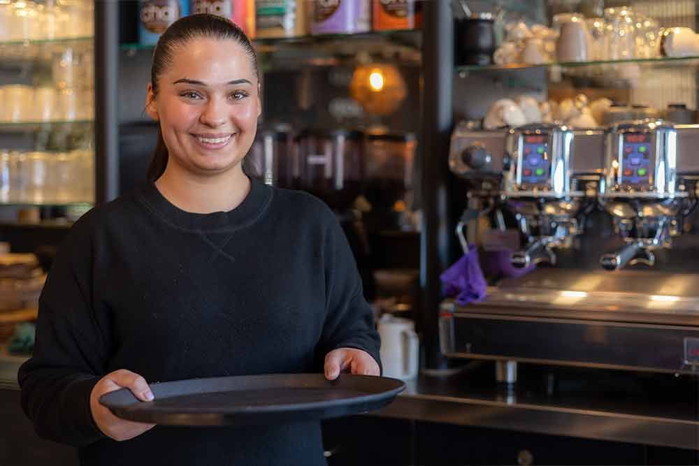 Eine lächelnde Frau hält ein Tablett in einem Café; im Hintergrund ist eine Espressomaschine zu sehen.