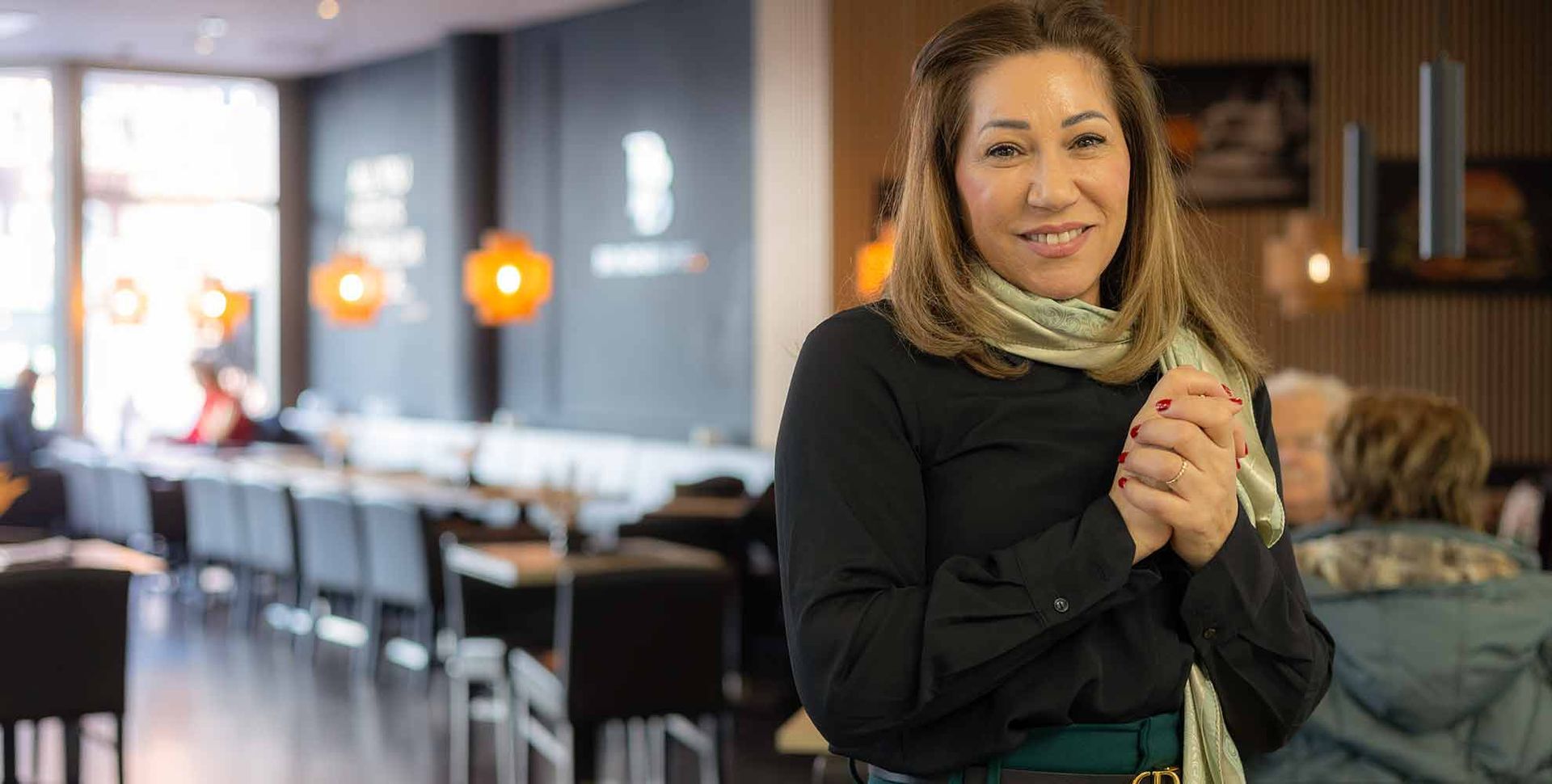 Frau lächelt in einem Restaurant, die Hände gefaltet, sie trägt einen Schal. Restaurantinterieur mit Tischen und Gästen.