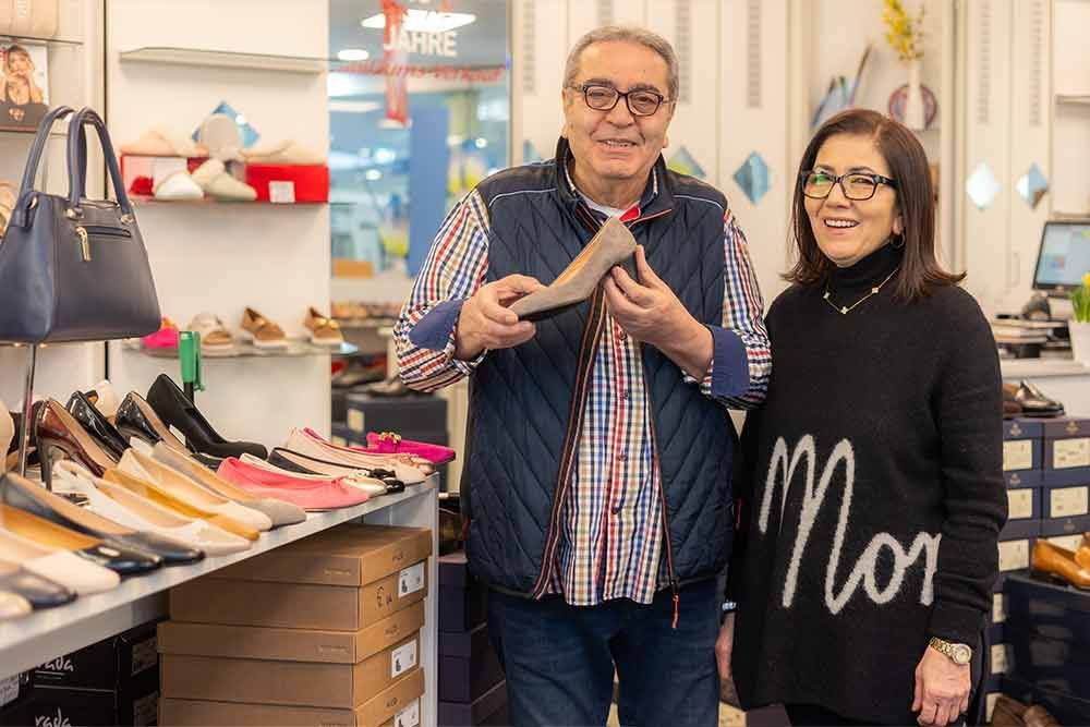 Ein Mann hält einen Schuh und steht mit einer Frau in einem Schuhgeschäft. Regale voller Schuhe sind zu sehen.