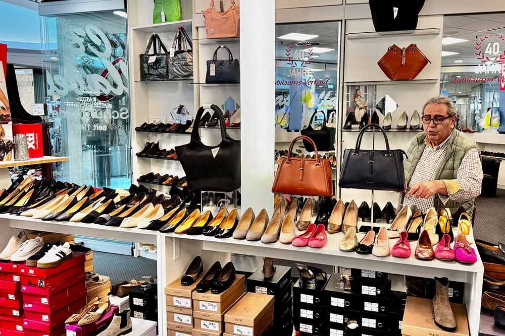 Ein Mann steht in einem Schuhgeschäft neben einer Auslage mit Schuhen und Handtaschen. Regale und Kartons sind mit Schuhen gefüllt.