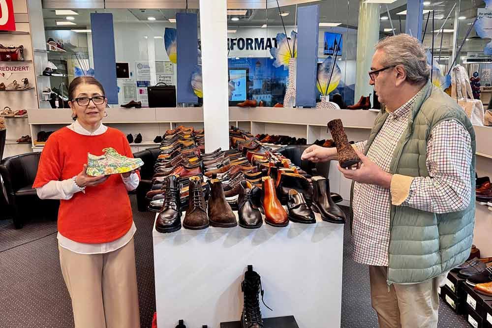 Frau hält Turnschuh, Mann hält Stiefel, beide in einem Schuhgeschäft, viele Schuhe ausgestellt.