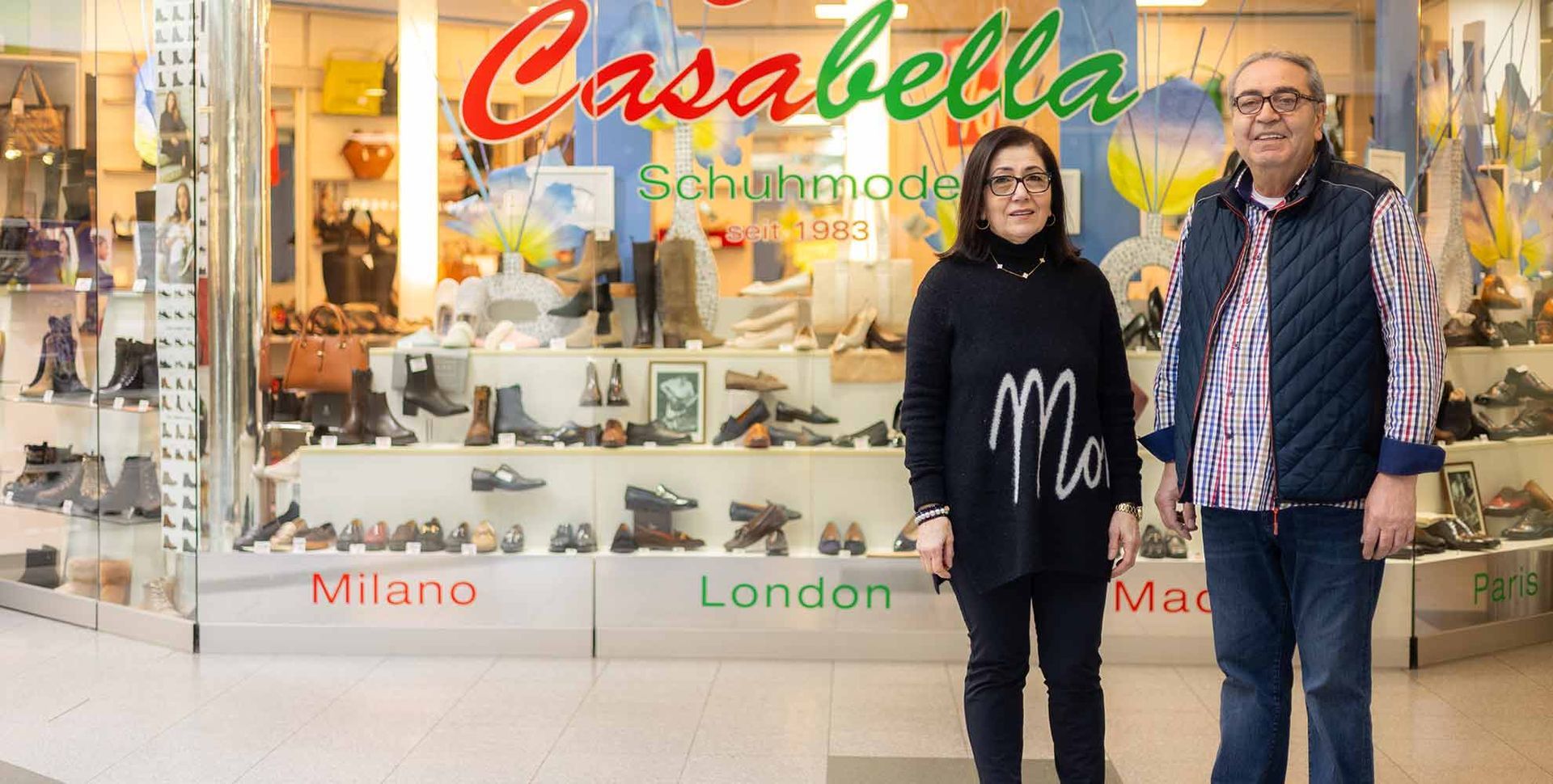 Zwei Personen stehen vor einem Schuhgeschäft namens Casabella. Im Schaufenster des Geschäfts werden verschiedene Schuhe ausgestellt.