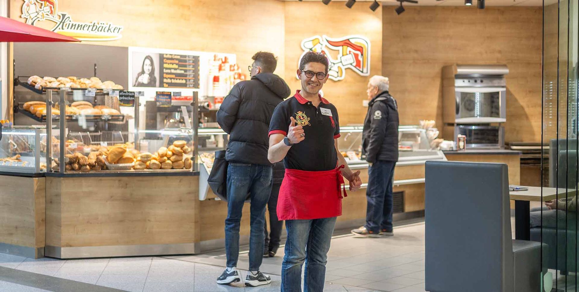 Bäckereimitarbeiterin mit roter Schürze zeigt den Daumen nach oben. Ladeninnenraum mit Backwaren und Kunden.