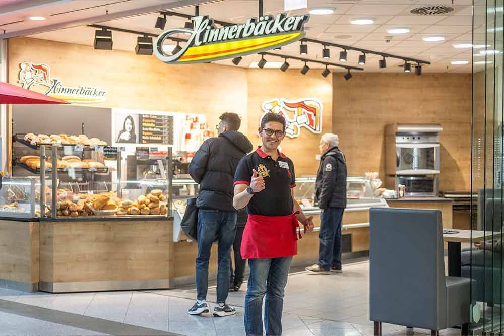 Eine Frau in roter Schürze lächelt vor einer Bäckerei namens „Kinnerbäcker“. Im Hintergrund sind Kunden zu sehen.
