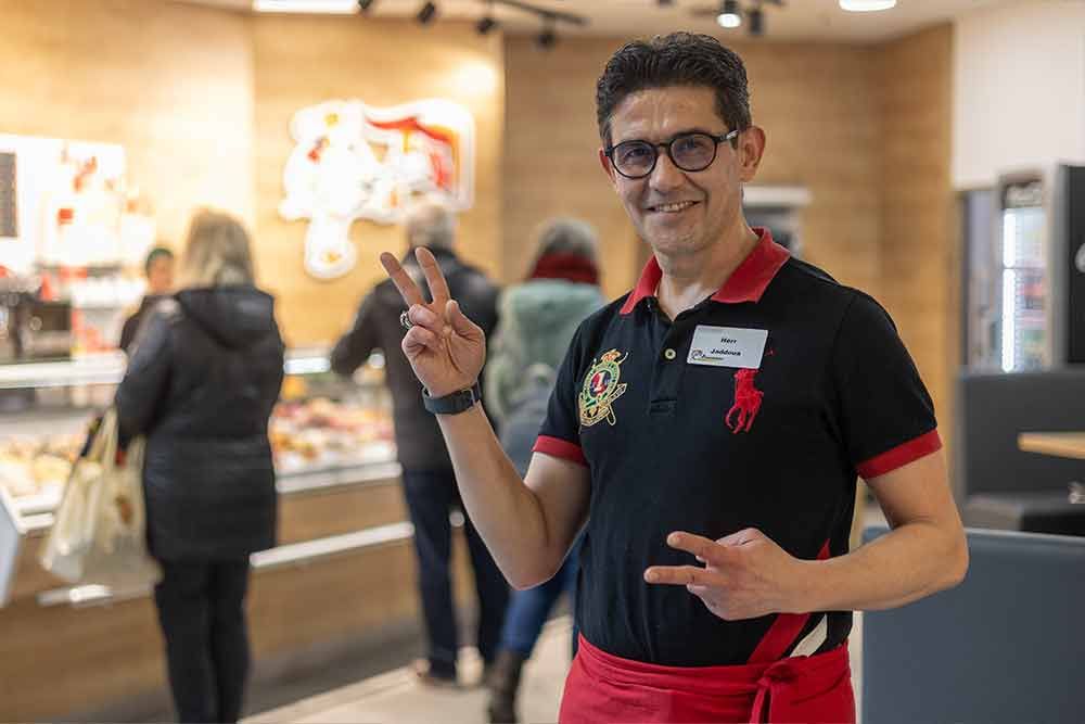 Ein lächelnder Mann in einer Metzgerei zeigt das Friedenszeichen. Er trägt eine rote Schürze und ein Poloshirt; im Hintergrund sind Kunden zu sehen.