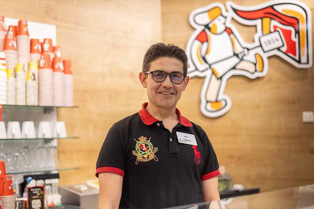 Ein Mann im Poloshirt lächelt vor einem Tresen. Im Hintergrund sind das Ladenlogo und Getränke zu sehen.