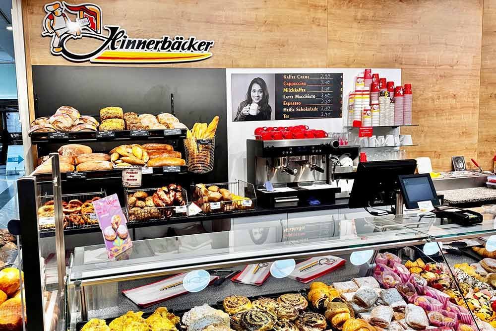 Auslage der Bäckereitheke: Gebäck, Brot, Kaffeemaschine, Frauenfoto, Schild „HinnerBäcker“.