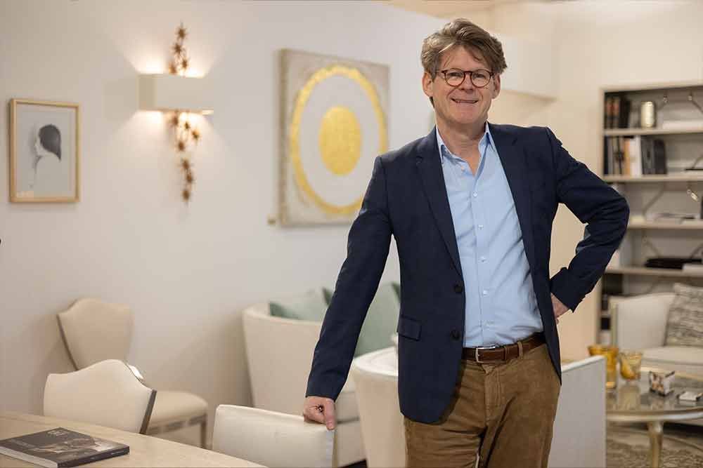 Ein Mann in blauer Jacke steht lächelnd in einem hell erleuchteten Wohnzimmer, die Hand auf einem Stuhl.