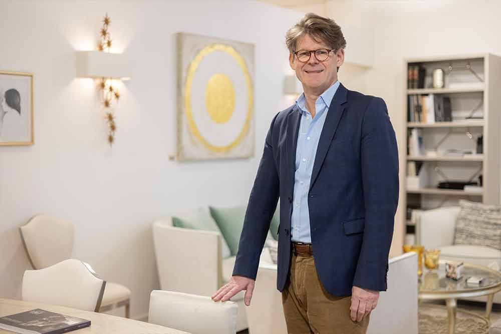 Ein Mann in Blazer und Anzughose steht in einem gut beleuchteten Wohnzimmer, die Hand auf einem weißen Stuhl, und schaut in die Kamera.