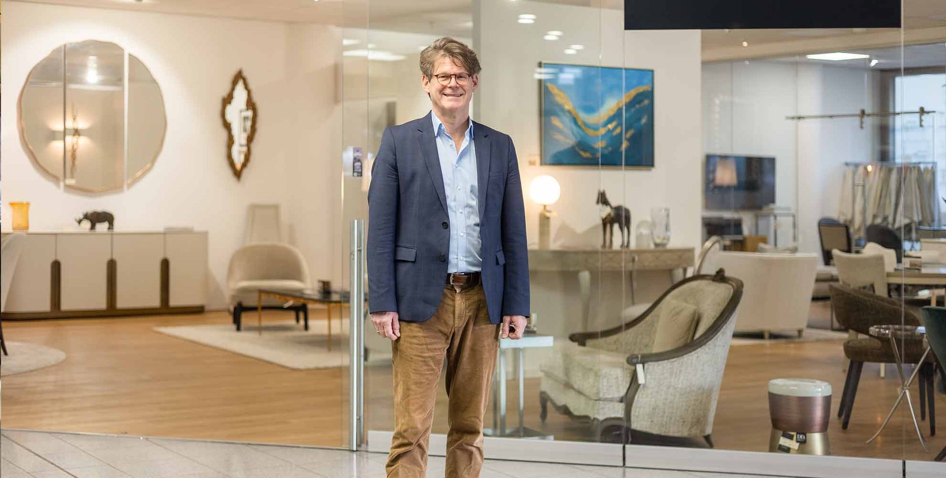 Ein Mann in Blazer und Cordhose steht lächelnd in einem Möbelgeschäft.