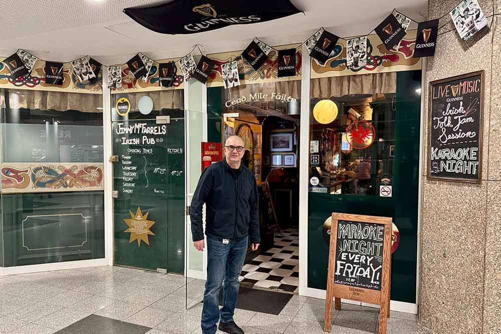 Ein Mann steht vor dem Eingang eines irischen Pubs mit schwarz-weiß kariertem Boden, Schildern und Dekorationen.