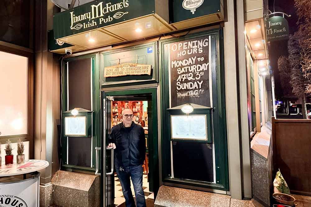 Ein Mann steht im Eingang eines irischen Pubs. Der Pub hat grüne Verzierungen und ein Schild mit den Öffnungszeiten.