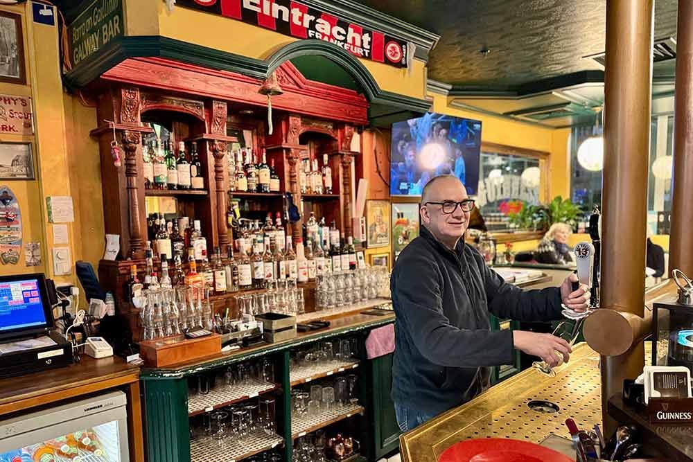 Ein Mann schenkt hinter einer Bar ein Getränk ein. Regale voller Spirituosen, Dekoration in Grün und Gold.