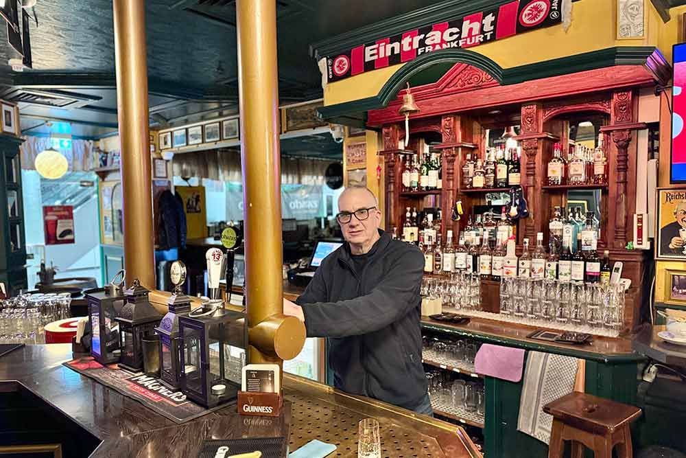 Mann hinter der Bar, Getränke in Regalen, Eintracht-Banner. Dunkles, klassisches Barinterieur.