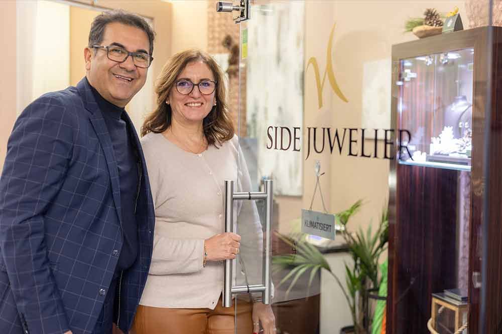 Zwei Personen stehen lächelnd im Eingang eines Juweliergeschäfts. Auf der Tür steht „SIDE JUWELIER“.