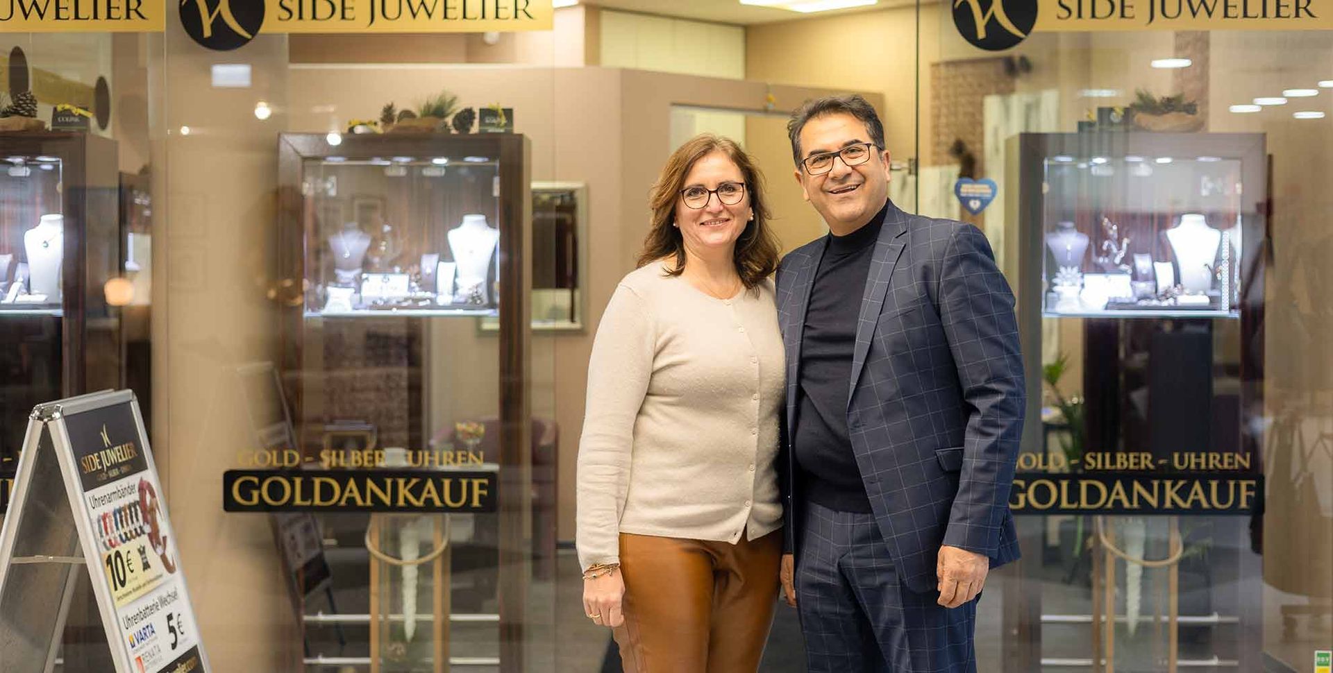 Zwei lächelnde Personen vor einem Juweliergeschäft mit der Aufschrift „Goldankauf“ im Schaufenster.