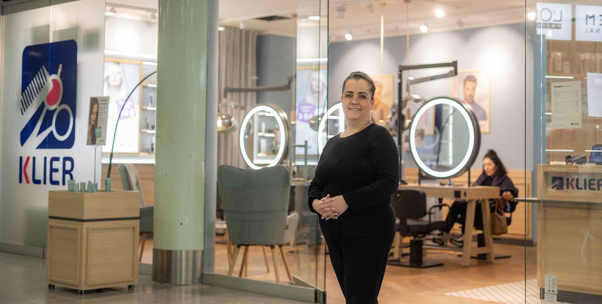 Eine Frau steht mit verschränkten Armen vor einem Klier-Friseursalon. Im Inneren sind eine Kundin und eine Friseurin zu sehen.