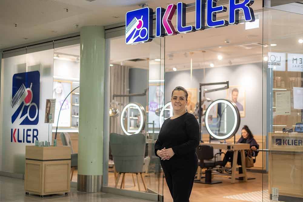 Eine Frau steht vor einem Klier-Friseursalon. Der Salon hat ein blau-weißes Schild, und eine Kundin befindet sich im Inneren.