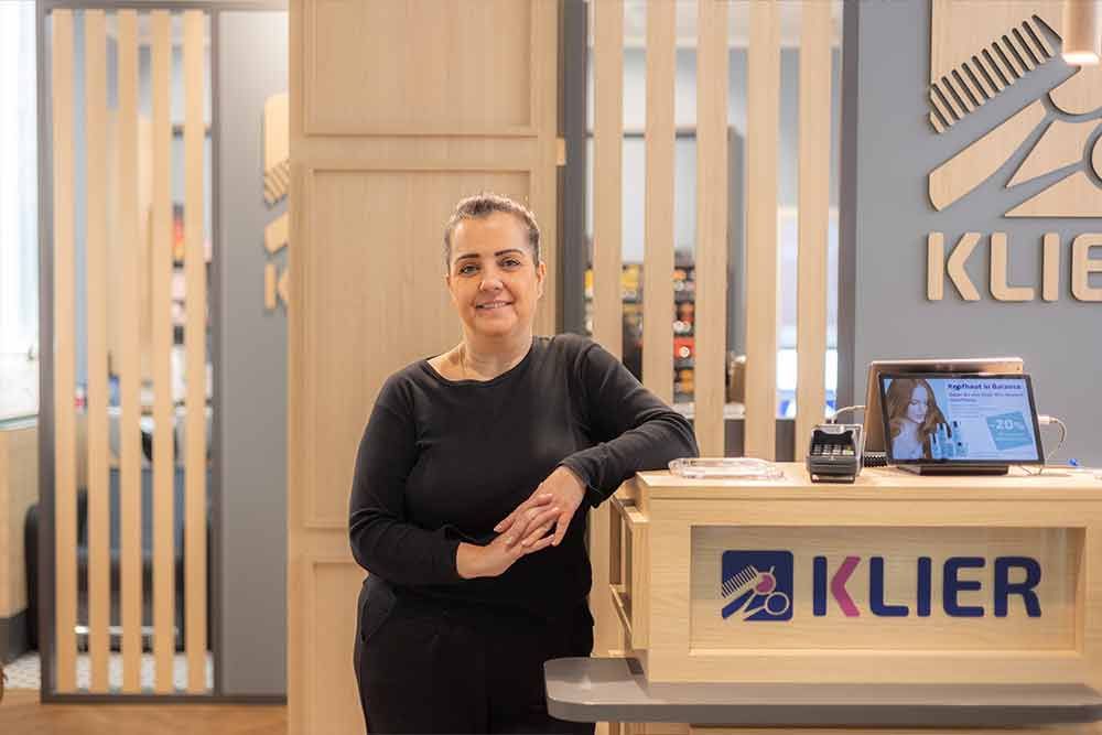 Eine Frau steht an der Rezeption eines Klier-Salons mit einem Schild und Logo vor einer hellen Holzvertäfelung.