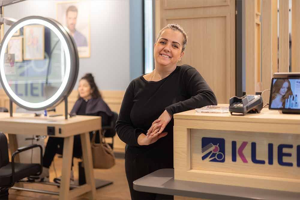 Eine Frau lächelt hinter dem Empfangstresen eines Salons mit dem Logo „Klier“; im Hintergrund sitzt eine Kundin.