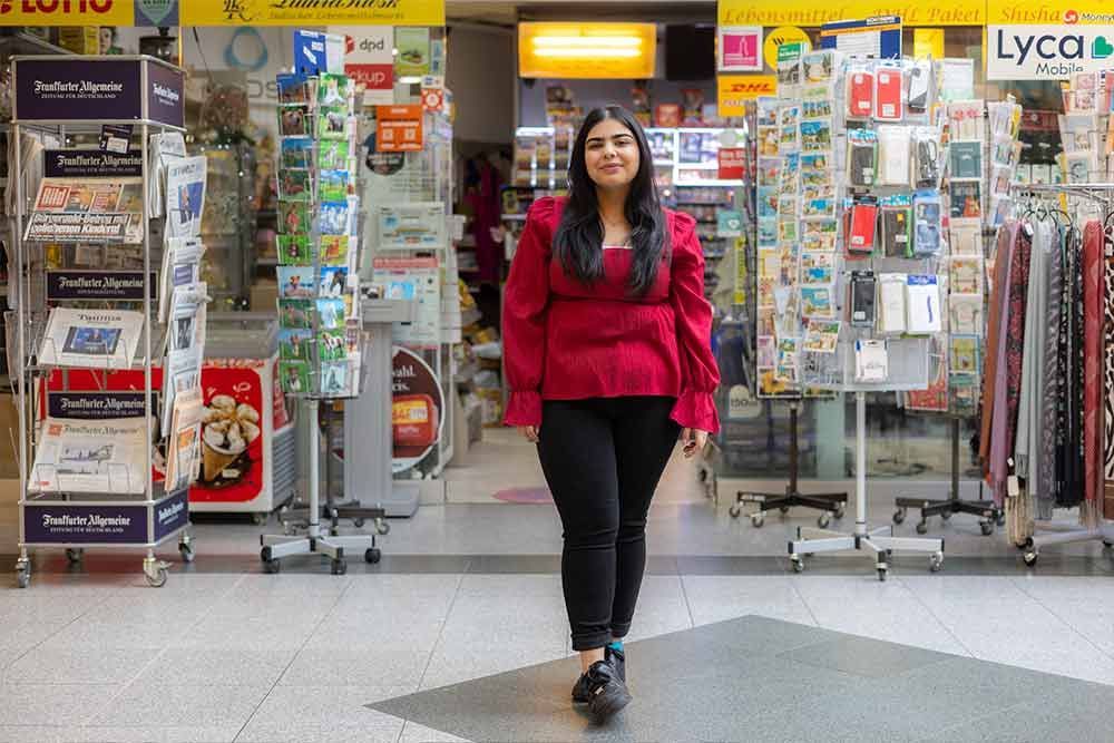 Frau in rotem Oberteil und schwarzer Hose steht im Eingang eines Geschäfts, umgeben von Waren.