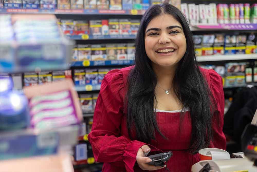 Eine Frau in einem roten Kleid lächelt an einer Kasse in einem Geschäft mit Regalen voller Produkte.