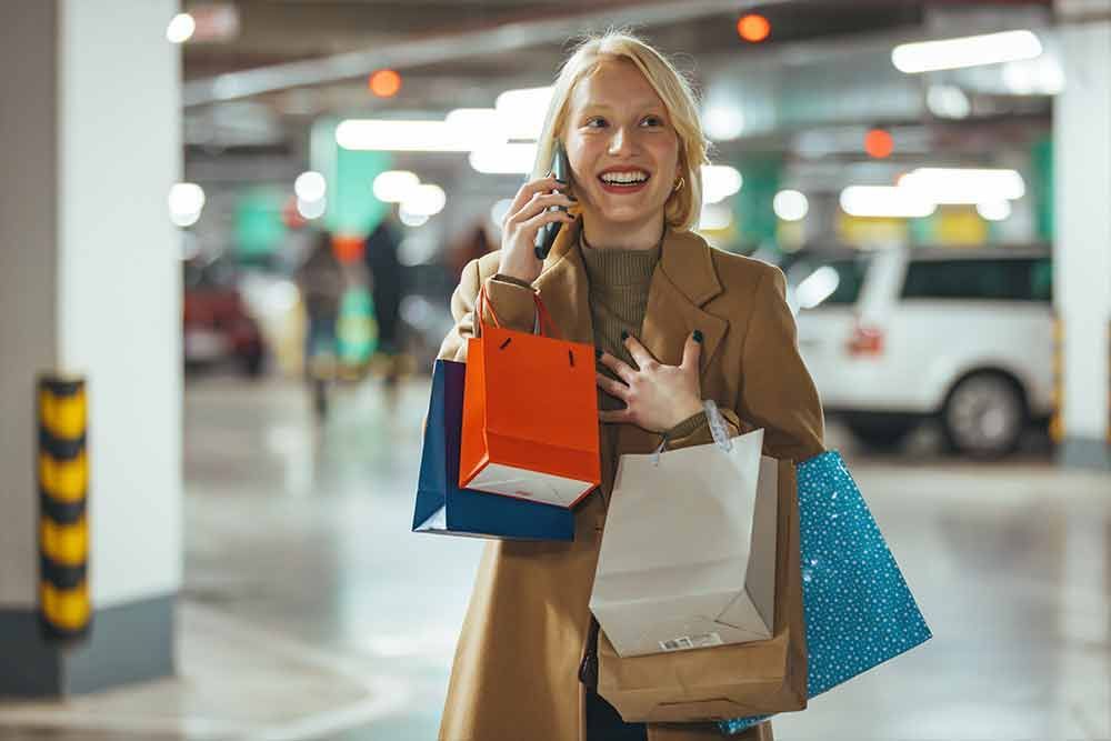 Frau im Mantel lächelt, hält Einkaufstüten und telefoniert in einem Parkhaus.