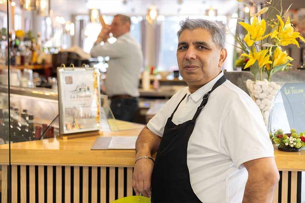 Ein Mann mit Schürze lehnt an einer Bartheke in einem Restaurant. Im Hintergrund ist ein weiterer Mann zu sehen. Gelbe Blumen sind erkennbar.