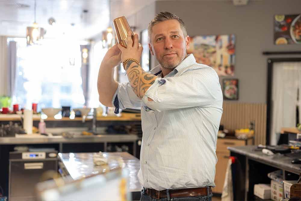 Barkeeper mit Tattoos am Arm, der in einer Bar einen Cocktailshaker schüttelt.