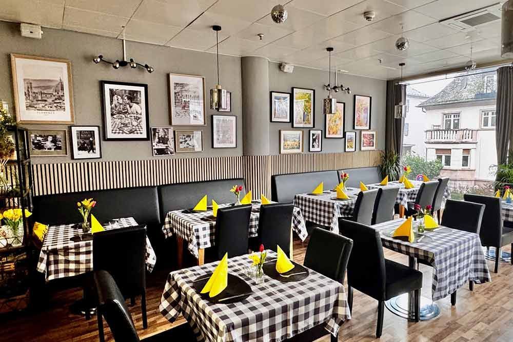 Restaurantinterieur mit karierten Tischdecken, schwarzen Stühlen und gerahmten Bildern an der Wand.